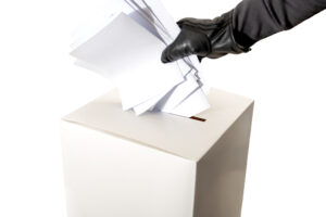 Electoral fraud and hacked elections concept with a hand wearing a leather glove and stuffing a ballot box isolated on white background with copy space and a clip path cutout